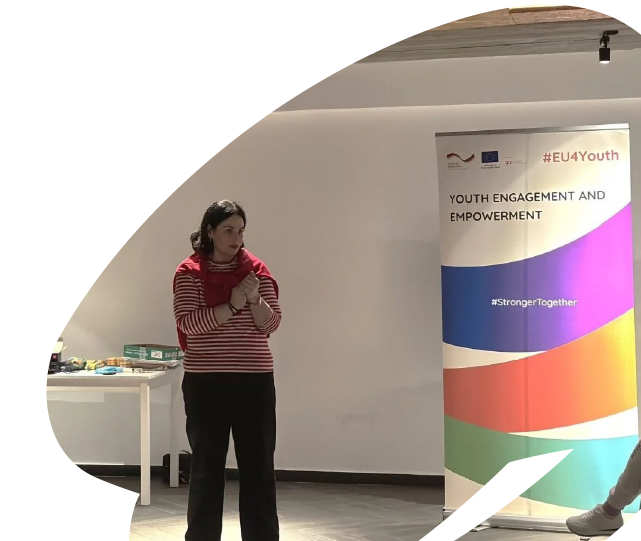 Facilitator speaking during an EU4Youth youth engagement session, standing beside an EU4Youth banner in a training or workshop setting. Facilitator speaking during an EU4Youth youth engagement session, standing beside an EU4Youth banner in a training or workshop setting.